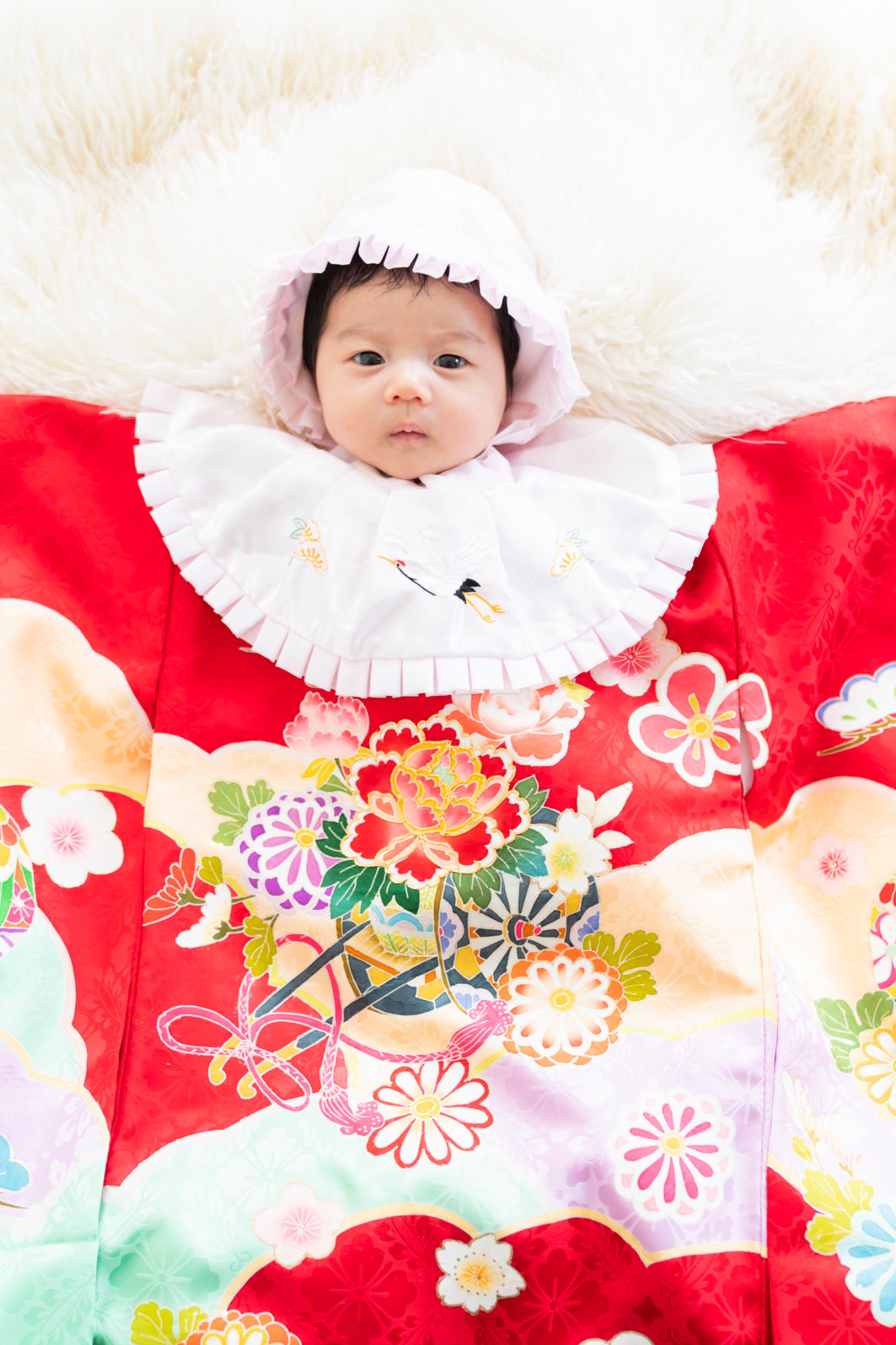 白い帽子と赤い産着姿で、こちらを見つめる赤ちゃん